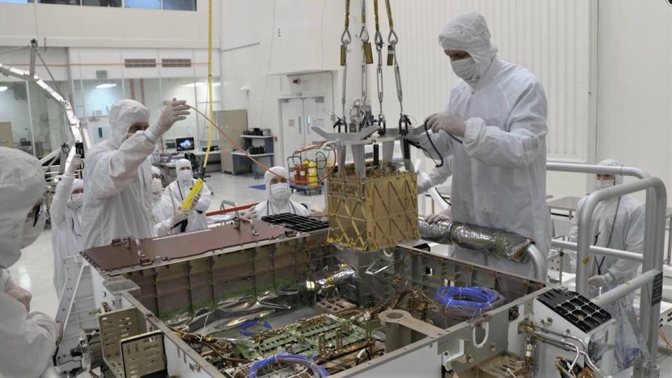 technicians in white clean suits lower a metallic box into a larger metal box