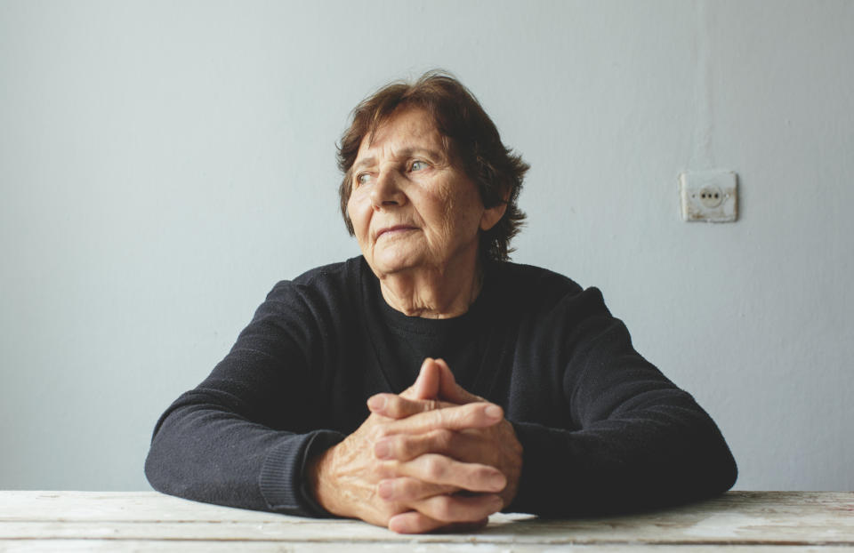 Elderly woman seated at a table, resting her chin on her clasped hands, looks thoughtfully to the side