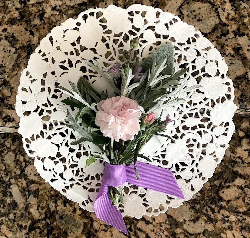 A fragrant tussie mussie in hues of pink, purple, and silver is ready to insert into a doily. The bouquet has a noisette rose bloom as its central blossom surrounded by fillers of lavender and sage blossoms and lamb’s ear, artemisia, and mint. Stems are gathered with a purple ribbon.