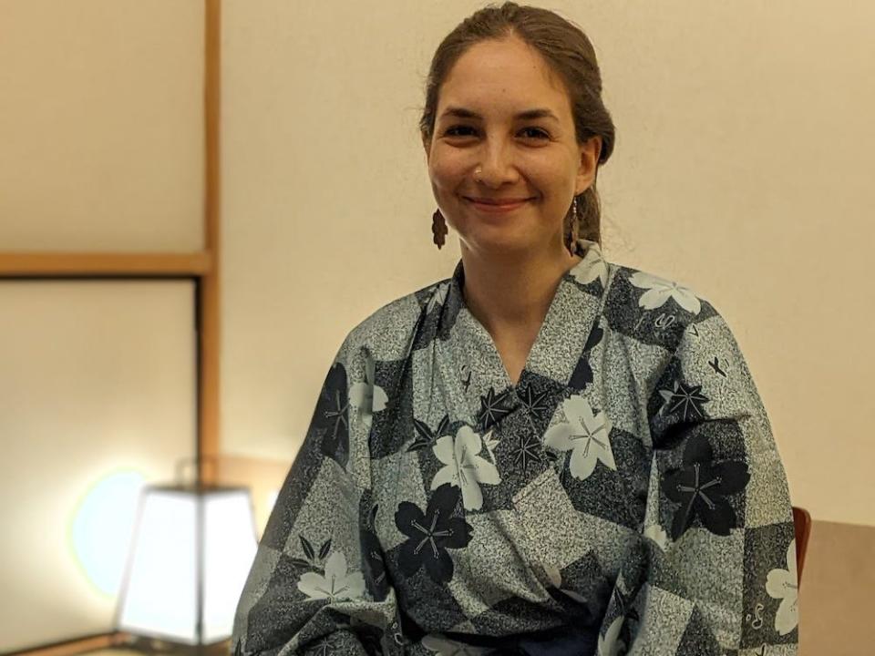 Photo of India Kushner sitting at a small wooden table wearing red earrings in the shape of a flower and a blue cotton robe with a floral abstract print. India has dark brown hair pulled into a ponytail and dark brown eyes. Behind her is a small lamp on the floor.