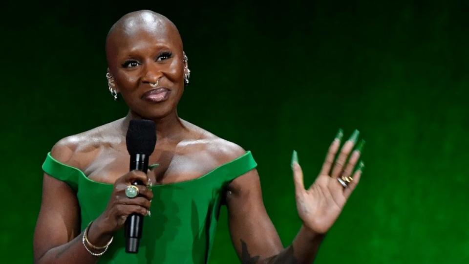 A Black woman stands holding a microphone and smiling on stage in front of a green background, holding up her hand with matching nail polish.