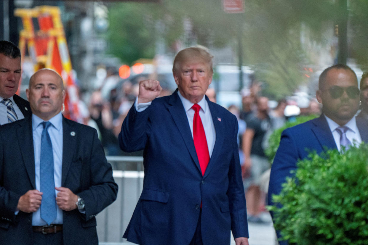 Donald Trump, holding up his right fist, is flanked by a few men in suits in front of a crowd of onlookers.