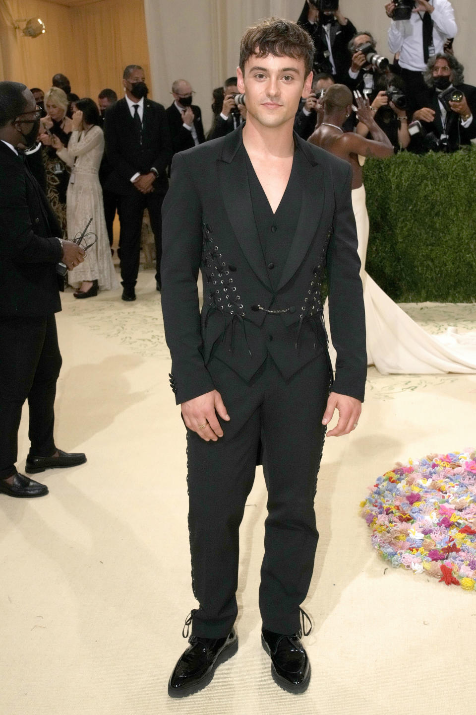 Tom Daley poses in a dark-colored suit with chains and lace-up details on the jacket