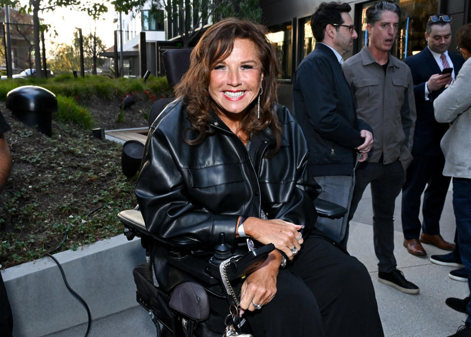 Abby in a leather jacket seated in wheelchair, smiling, with people standing in the background