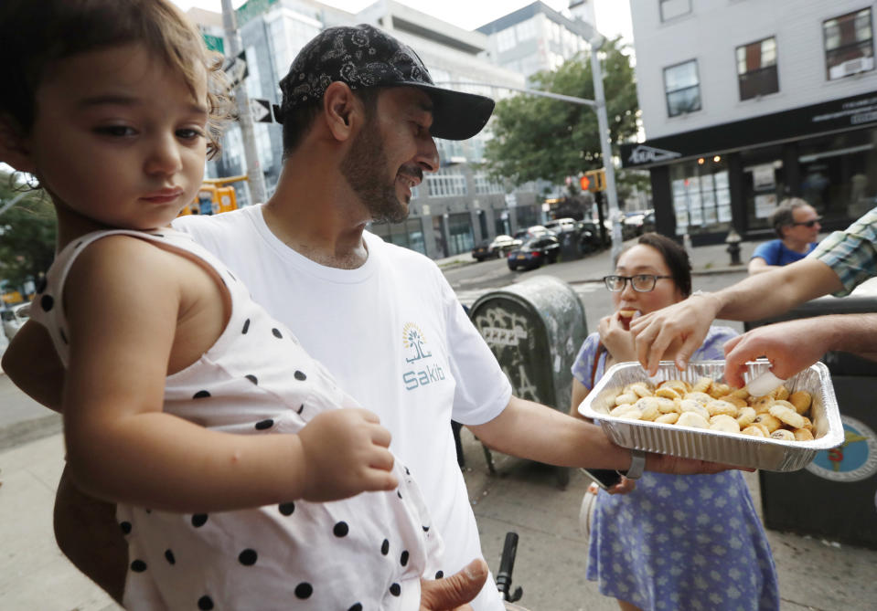 In this July 11, 2019, photo, Syrian refugee and restaurant owner Diaa Alhanoun holds his young daughter, Masa, then 21 months, while offering his wife's homemade cookies to passers-by outside his restaurant, Sakib, on it's opening night, in the Williamsburg section of Brooklyn in New York. Of all the places he’s lived, Alhanoun thinks New Yorkers have liked his food the most, and he’s hopeful about what the future could hold as he builds a new life here. (AP Photo/Kathy Willens)