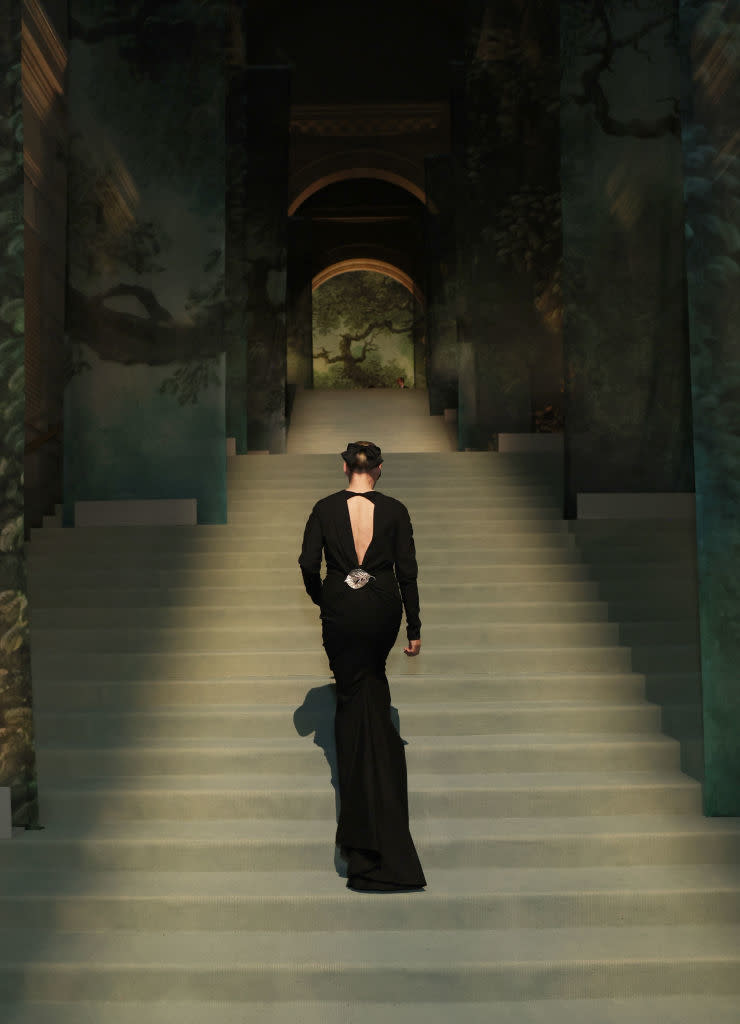 Individual in an elegant backless gown ascending a grand staircase with painted walls