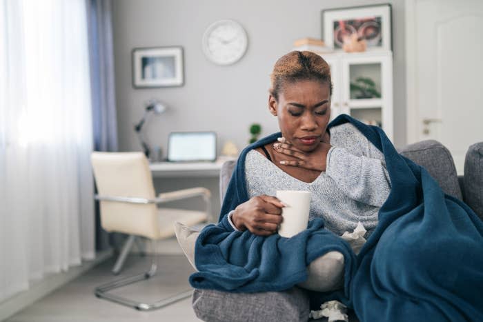 Person holding a cup while sitting on a couch covered in a blanket, appearing to feel unwell, in a home office setting. No other identifiable persons