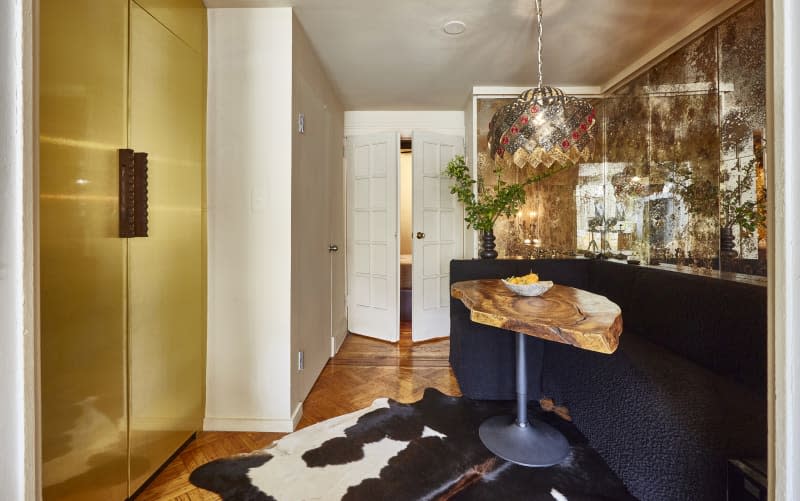 dining area in apartment with antiqued mirror, rustic wood table, black banquette, hide rug. Closet with gold doors across the hall