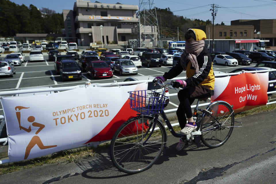 Una mujer pedalea frente a carteles con el logo del relevo de la antorcha olímpica en Naraha, Japón, el miércoles 24 de marzo de 2021. (AP Foto/Eugene Hoshiko)