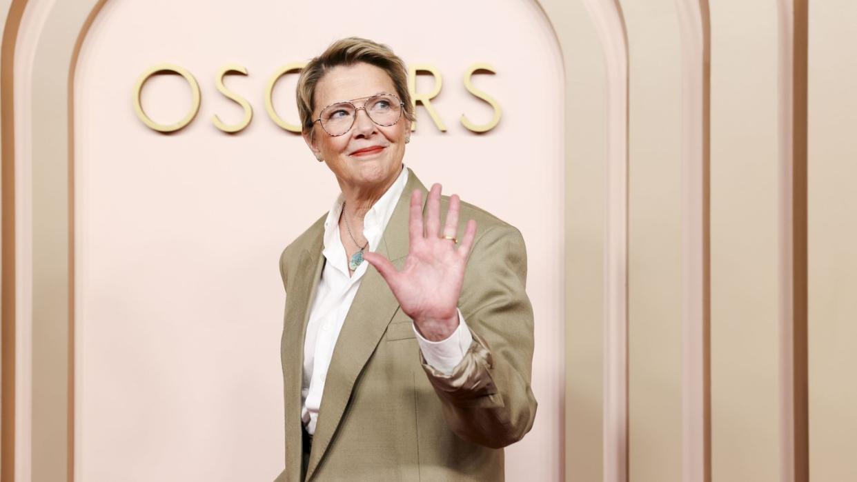 annette bening smiles and waves while walking in front of a wall with an oscars logo, she looks to the right and wears a tan suit jacket, white collared shirt, glasses, and a necklace