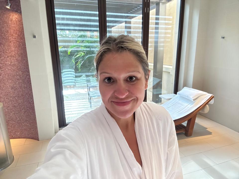 A selfie of Terri Peters in a white robe at a spa. Her blond hair is pulled back. She has a nose stud in her left nostril, and she smiles slightly. Behind her is red tiled wall, a floor-to-ceiling glass window, and a lounge chair with a white towel on top.
