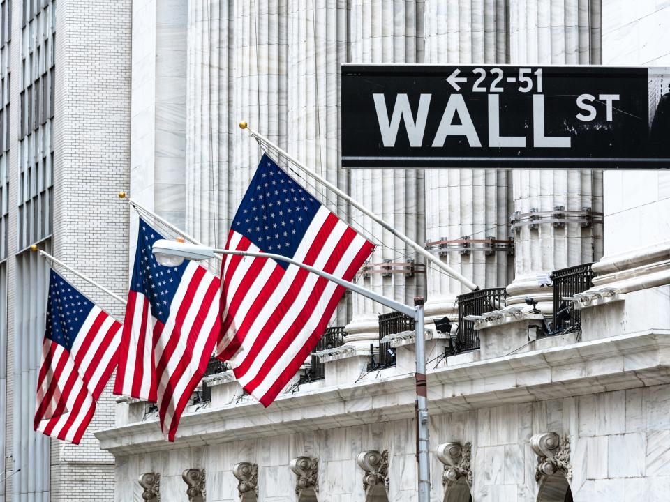 A Wall Street sign in front of the New York Stock Exchange, which has three American flags on its facade.