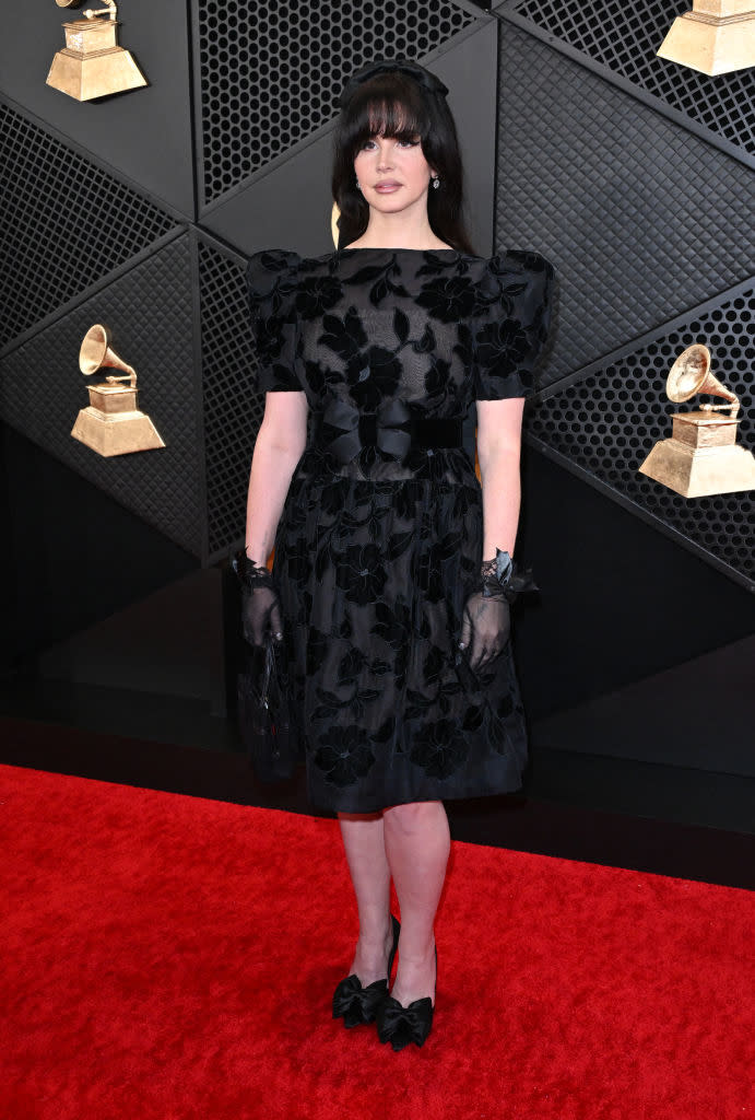 Woman in black, floral-patterned dress and gloves stands on red carpet; Grammy Awards in background