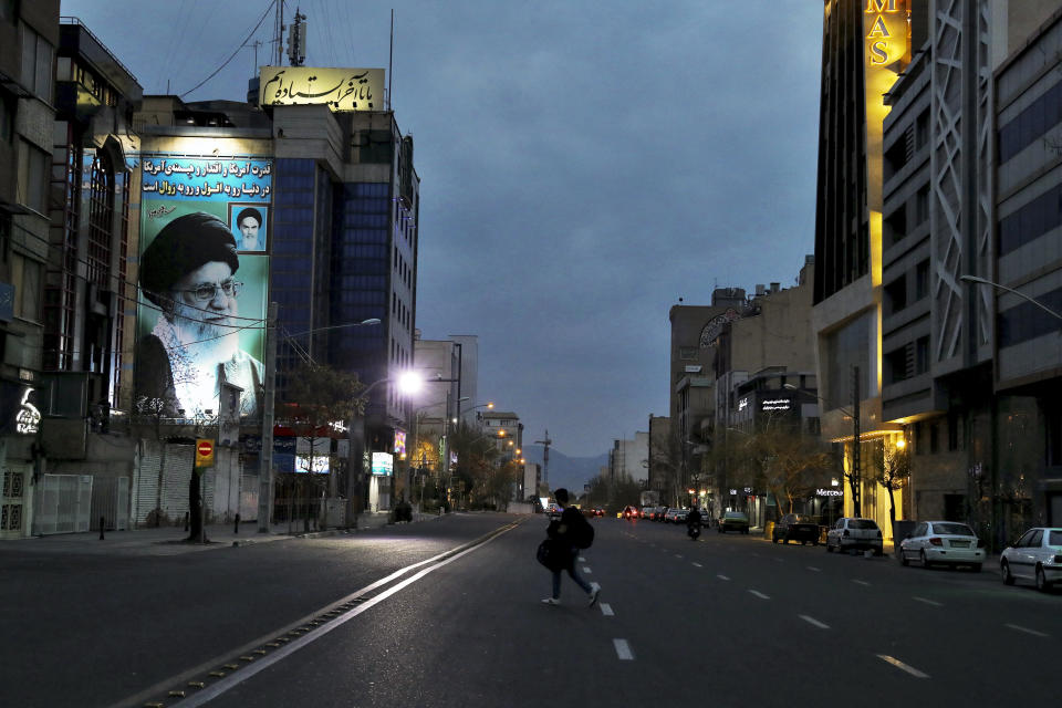 A pedestrian crosses an empty street in Tehran, Iran, on Tuesday, March 24, 2020, with a portrait of Iran's Supreme Leader Ayatollah Ali Khamenei at right. Due to the coronavirus and the COVID-19 illness it causes, life has changed dramatically on Tehran's main highways, in the narrow corridors of its historical Grand Bazaar and in the public spaces under portraits of its leaders. (AP Photo/Ebrahim Noroozi)