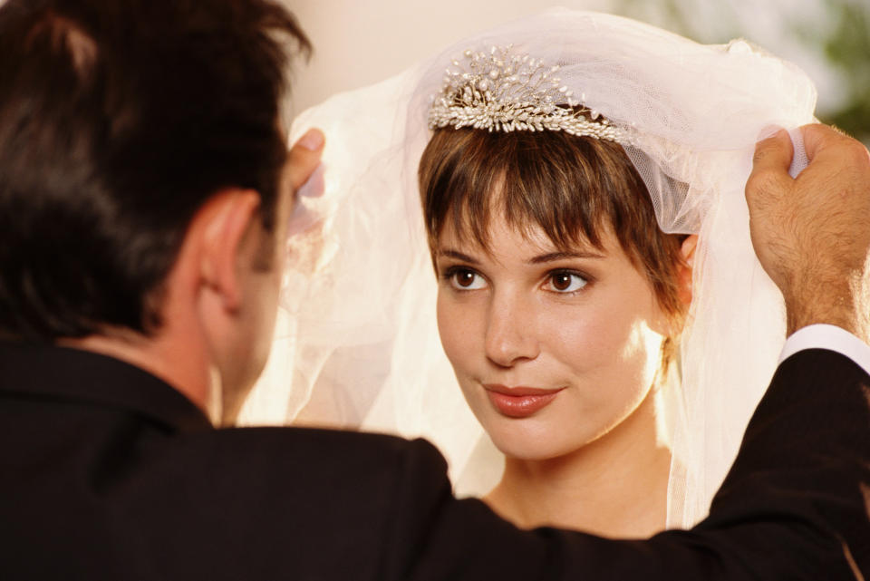 A bride in a wedding dress and veil, assisted by an unidentified man in a suit. The veil features a decorative headpiece