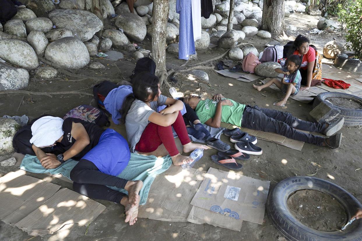 Children and adults sit and lie on the bare dirt ground amid broken-down cardboard boxes and cut-up tires.