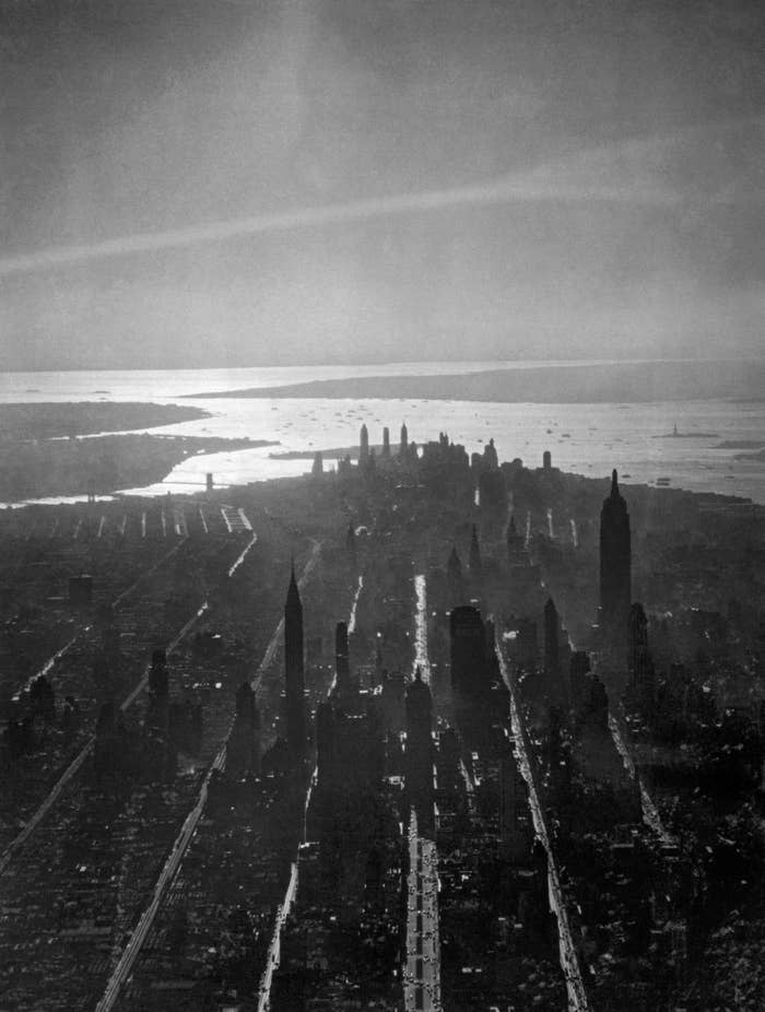 Aerial view of New York City with the skyline and skyscrapers casting long shadows. The horizon features large water bodies in the distance