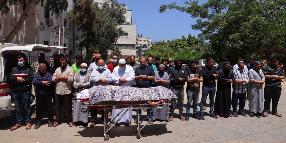 A group of men outdoors in Gaza standing in prayer pose behind a trolley holding the shrouded body of a Palestinian.