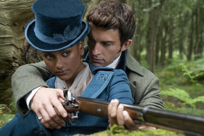 a man stands behind a woman and helps her aim a shotgun