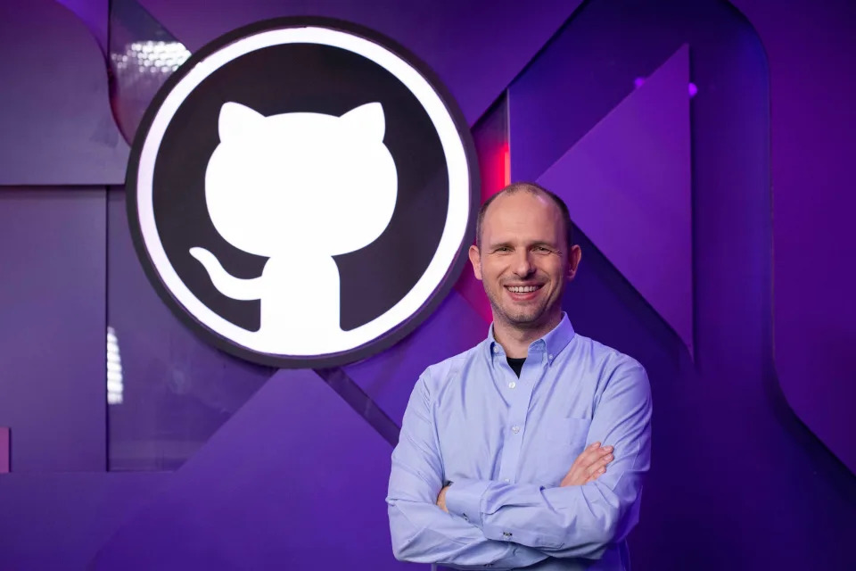 Thomas Dohmke standing cross-armed in front of the GitHub logo.