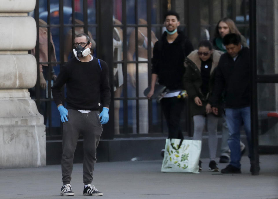 A man wearing a mask waits with other people at a bus stop in London, Tuesday, March 24, 2020. Britain's Prime Minister Boris Johnson on Monday imposed its most draconian peacetime restrictions due to the spread of the coronavirus on businesses and public gatherings. The highly contagious COVID-19 coronavirus can cause mild symptoms, but for some it can cause severe illness including pneumonia.(AP Photo/Frank Augstein)