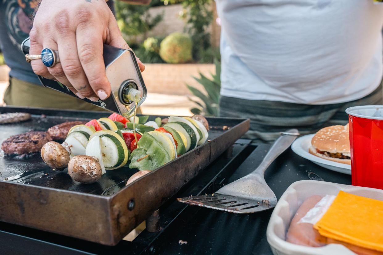 Close-up shot of a man dousing cooking oil on a skewer with vegetables; bell peppers, zucchini, mushroom, onion, and tomatoes, atop a grill.