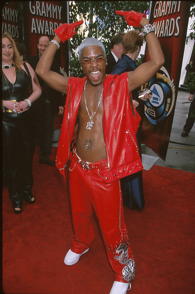 Sisqo wearing a leather vest and pants and smiling with his hands up in the air on the red carpet