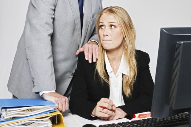 A  businessman stands behind a pretty and vulnerable young businesswoman, leaning over her with one hand on her shoulder as she