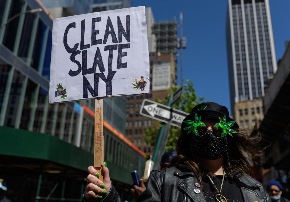 Below a skyscraper, a demonstrator wearing green nail polish and with green leaves on their eyes marches in the parade with a placard that says Clean Slate NY..