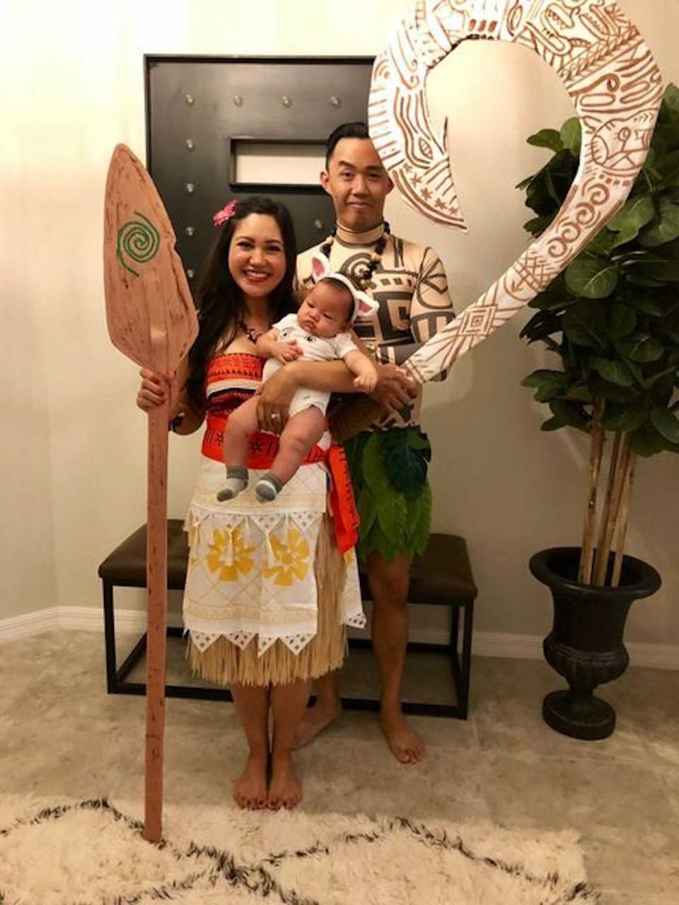 A couple dressed as Moana and Maui pose holding a paddle, sword, and a baby.