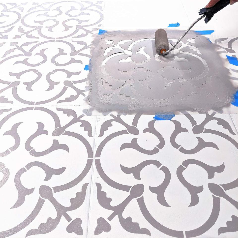 Using a small paint roller to apply grey paint onto a tile floor, using a stencil. 