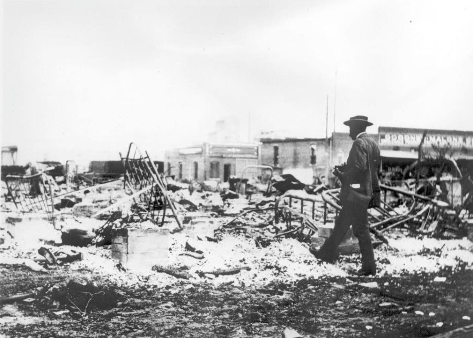 Black-and-white photo of a burned landscape, with some still-standing buildings in the background, as one man walks on the rubble