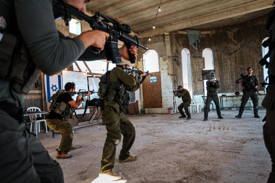 People in fatigues stand and point assault weapons at others facing them inside a building