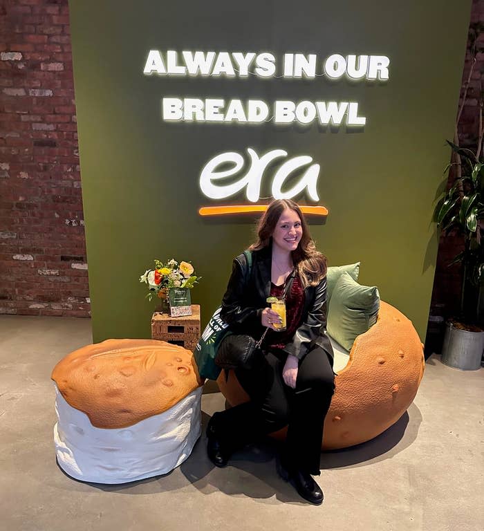 Woman sitting on a bread bowl-shaped chair at an "ALWAYS IN OUR BREAD BOWL ERA" event