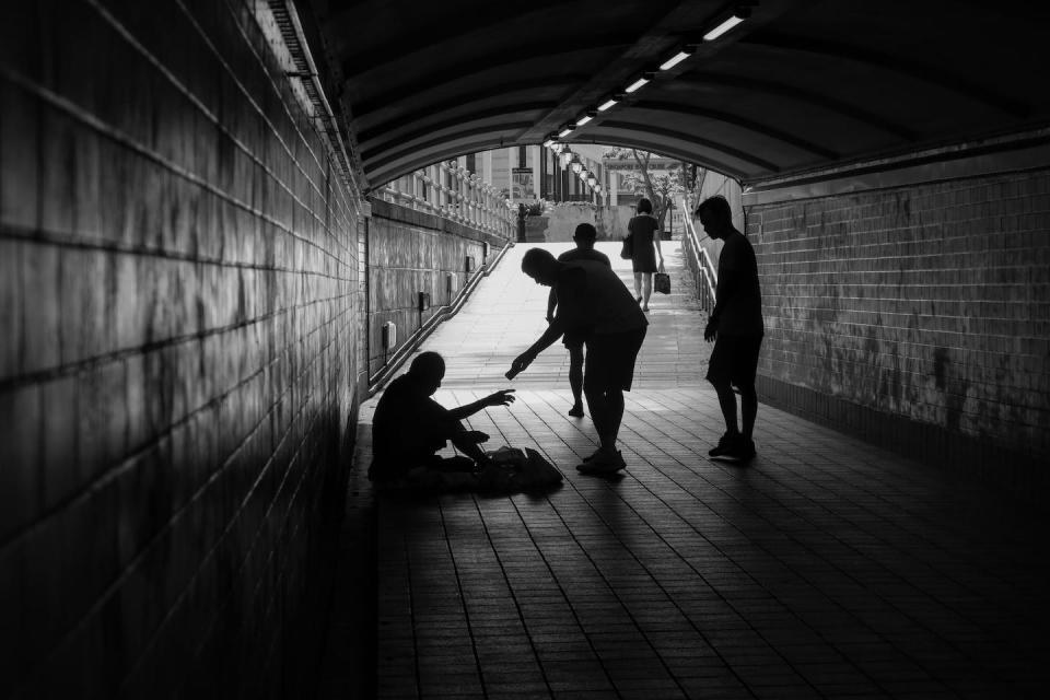 One person stops to give something to another person sitting on the ground in a tunnel.