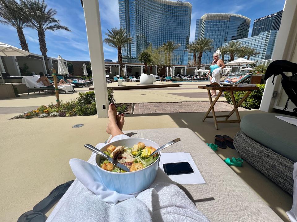 Someone sitting on a pool chair in a towel with legs visible and a salad on their lap.