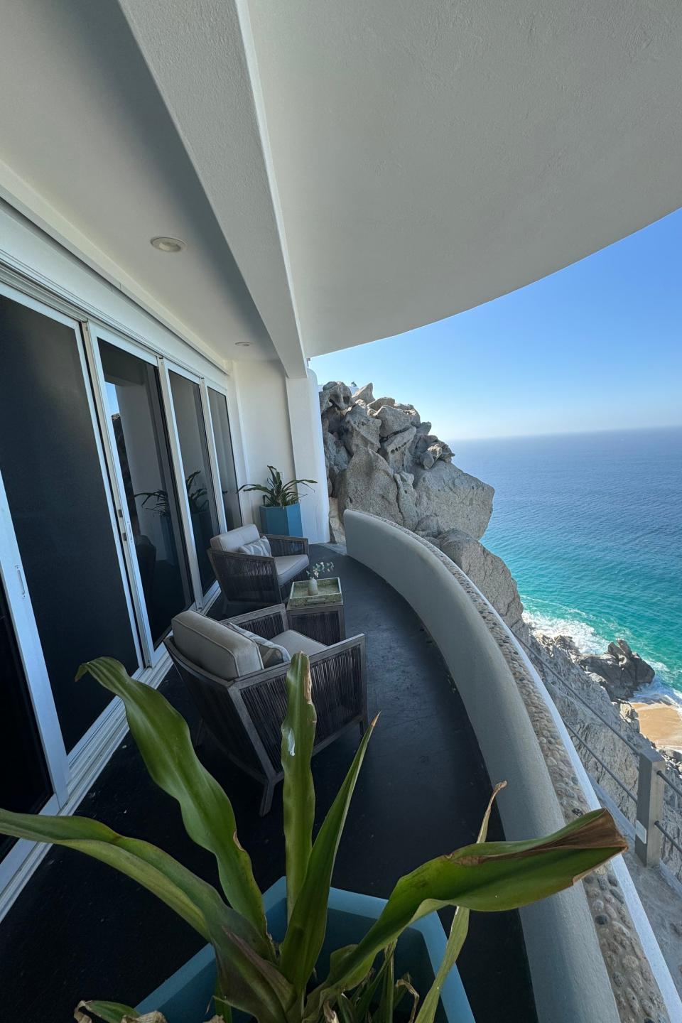 Balcony view overlooking the ocean from a coastal cliffside. Two lounge chairs face the sea, next to a small table and a potted plant, providing a serene retreat