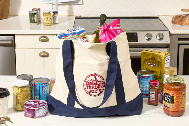 angled shot of an assortment of trader joe's groceries in a khaki and blue trader joe's tote bag - and some of the groceries are scattered around the bag on the counter.