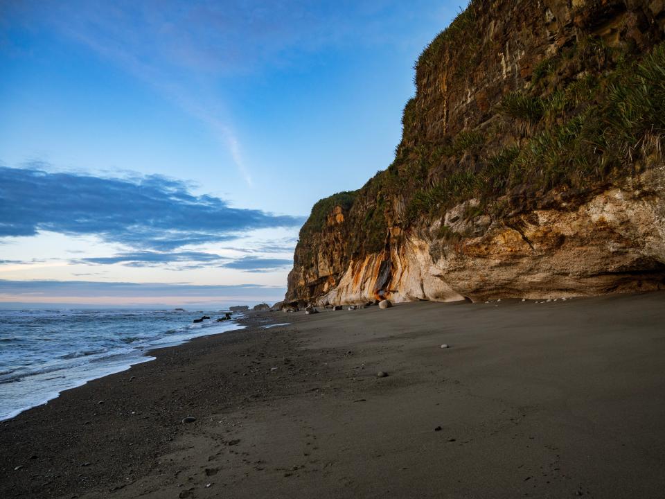 A rocky bluff in front of a black sand beach at sunset.