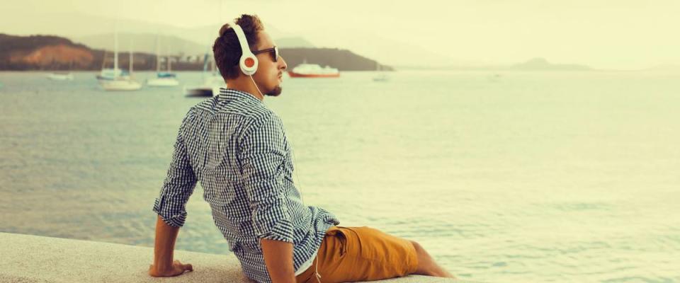 stylish young man in a shirt and shorts and sneakers listening to music in headphones on a beach