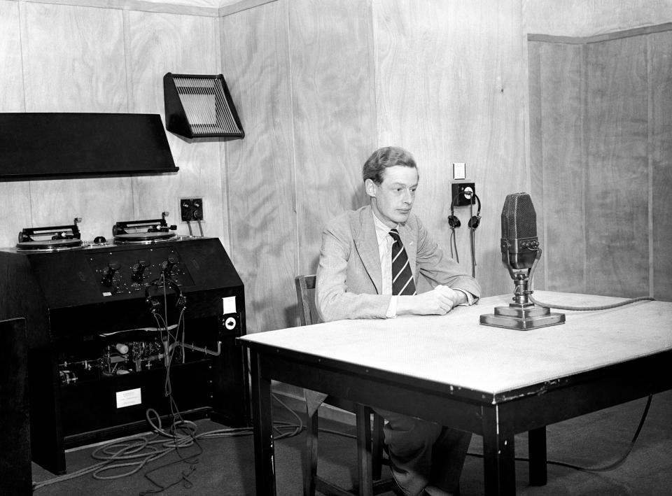 Announcer in a studio with a microphone in front of him and gramophone reproducing equipment in the background.