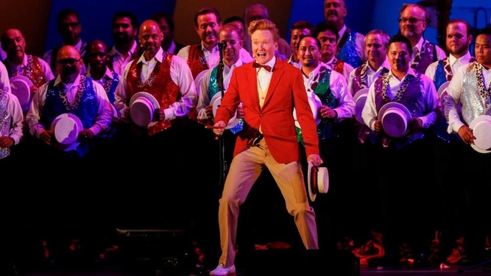 A man in a red jacket and bowtie sings loudly in front of a group of men singing in colorful vests, on stage.