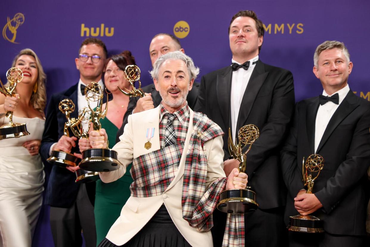 Alan Cumming stands in front of a group of people, all holding Emmys