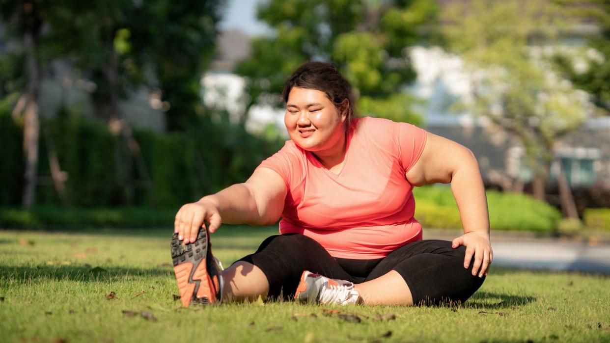 asian overweight woman exercising stretch alone in public park in village, happy and smile in morning during sunlight fat women take care of health and want to lose weight concept