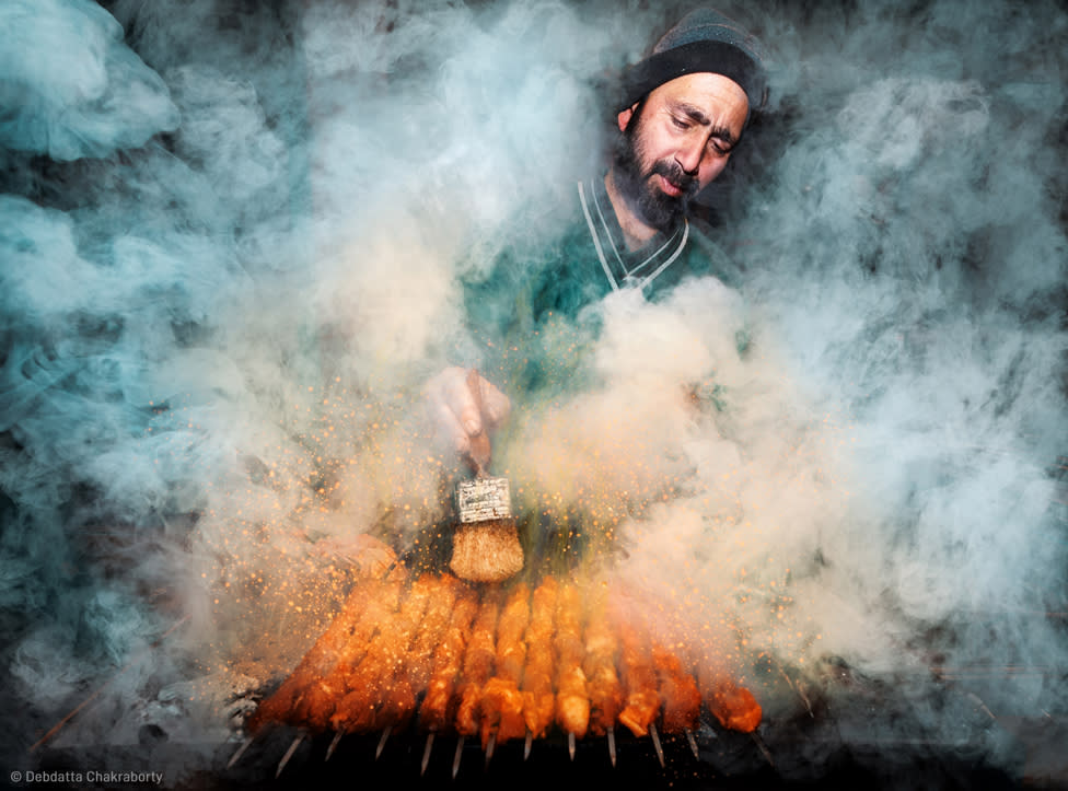 Vendedor de comida en la calle rodeado de humo mientras prepara unas brochetas.