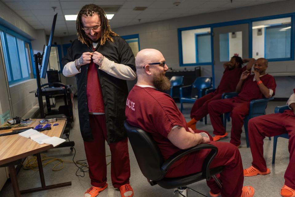 James Wheeler, center, sits in a barber chair as Eryn Carver, left, puts on a barber jacket before cutting his beard during a one-hour barber course for a small group of incarcerated men as part of the I.G.N.I.T.E. (Inmate Growth Naturally and Intentionally Through Education) program held in an activities room of the Genesee County Jail in Flint on Thursday, Jan. 4, 2024. Carver invited Wheeler to sit in for the class as he expressed wanting to get his beard cut before an upcoming court date.