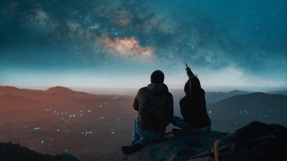     Two people sit together and look at the night sky with their naked eyes. The person on the right points at the stars. 