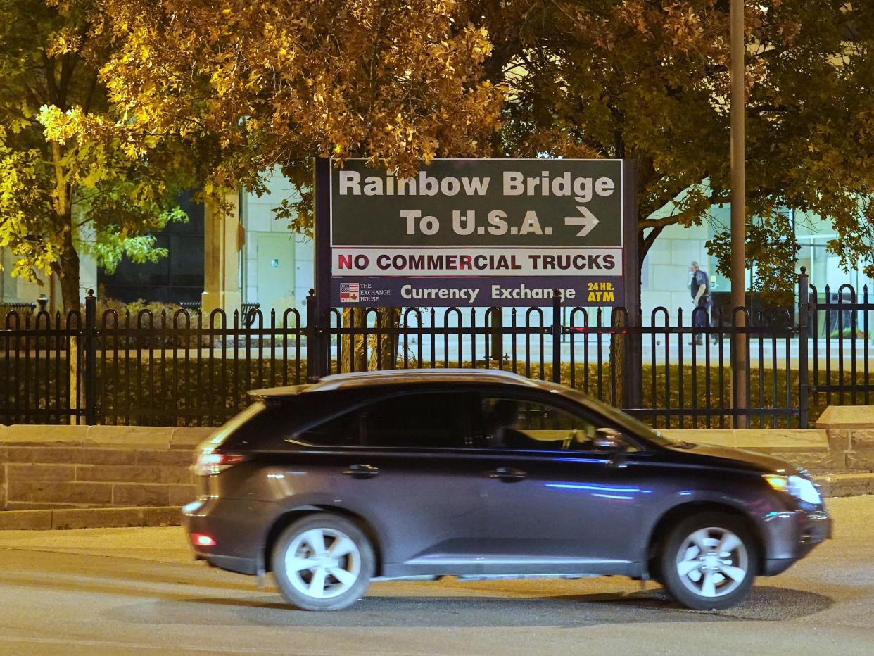 A gray four-door car driving down a road at night next to a group of trees and a sign which reads "Rainbow Bridge To U.S.A."