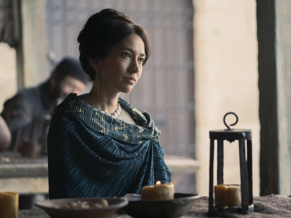 sonoya mizuno as mysaria in house of the dragon. she's a relatively young woman, wearing a blue shawl and with neat hair, sitting at an outdoor tavern table