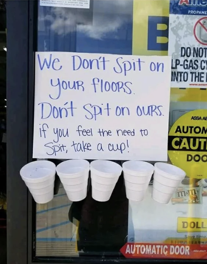 The sign says "We don't spit on your floors, don't spit on ours; if you feel the need to spit, take a cup" with a row of five cups taped to the bottom of the sign